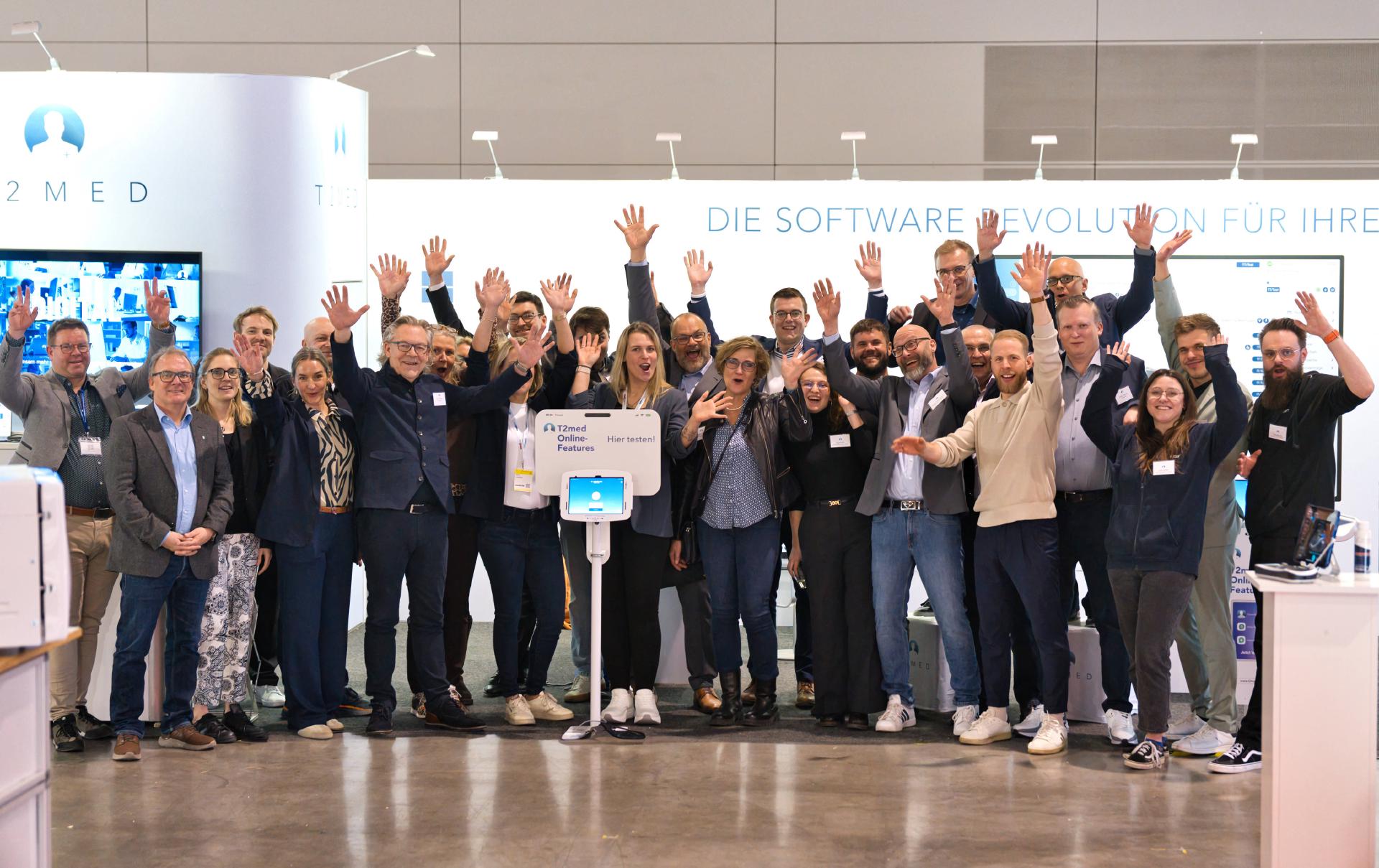 T2med-Mitarbeiter:innen und Partner:innen stehen jubelnd vor dem T2med-Messestand